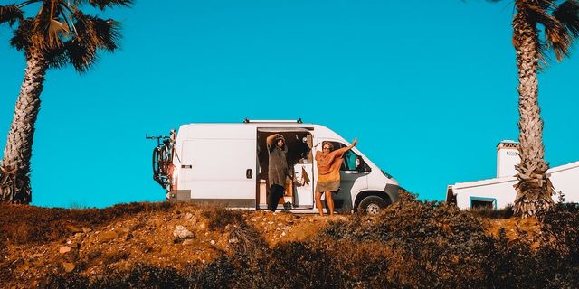 Two people standing in front of a parked CamperBoys van, the sky is blue and the sun is shining.