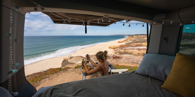 Man sieht aus dem Innern eines Campers eine Frau neben dem Auto am Strand sitzen und Essen zubereiten. Bayerische Karibik und Karibik in Europa - wir stellen dir die schönsten Spots vor.