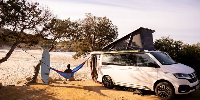 A parked CamperBoys van with a person sitting in a hammock next to it and a surfboard leaned on a tree.