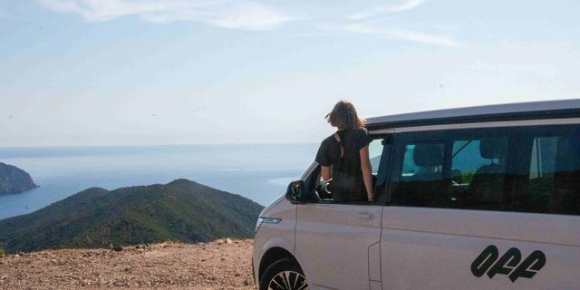 Woman looks out of window of off camper onto the sea in Corsica