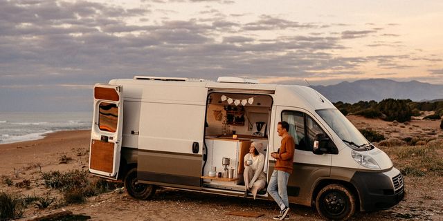 A woman sitting in a parked CamperBoys van and a man outside leaning against the van. They are both drinking something and  looking happily into the same direction.
