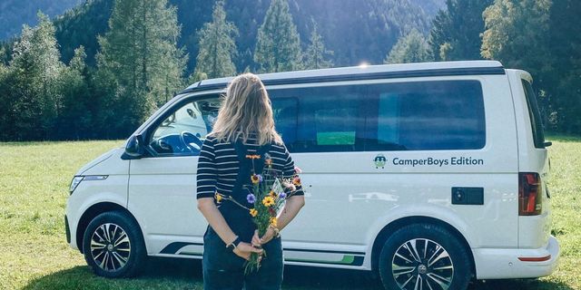 Mädchen steht vor VW California Beach auf einer Wiese und hält einen Wildblumenstrauß hinter dem Rücken