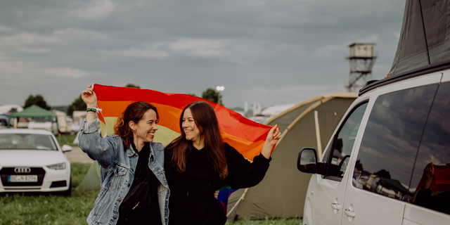 Lgbt freundliche Reiseziele fürs Campen: Ein lesbisches Pärchen auf einem Campingplatz hält eine Regenbogen-Flagge in den Händen und lacht sich an