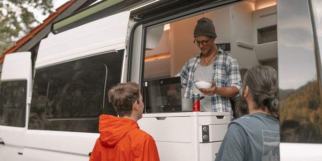 One man cooking inside the camper van and another man standing outside the camper van 