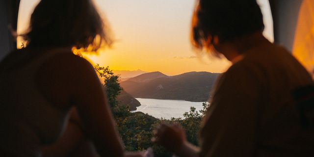 Zwei Frauen sitzen hinter einem Camper Van und betrachten den Sonnenuntergang.