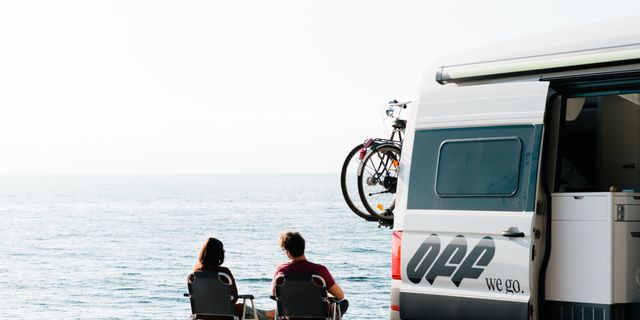 Ein Mann und eine Frau sitzen auf Campingstühlen neben einem Camper, an dem ein Fahrradträger montiert ist