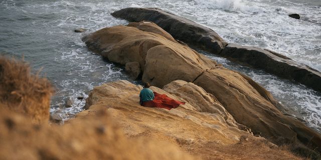 Eine Person sitzt mit einer Decke auf einem Felsen an der Küste und blickt aufs Meer.