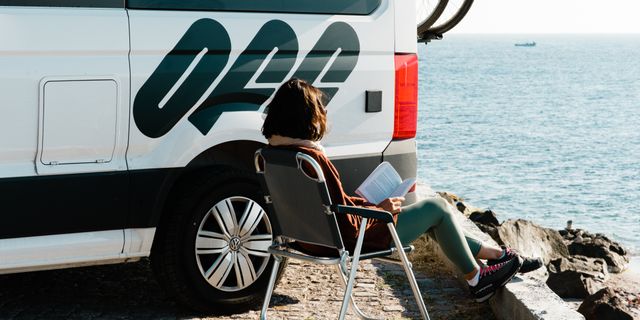 Ein Off Camper steht vor dem Meer, daneben liest eine Frau im Campingstuhl