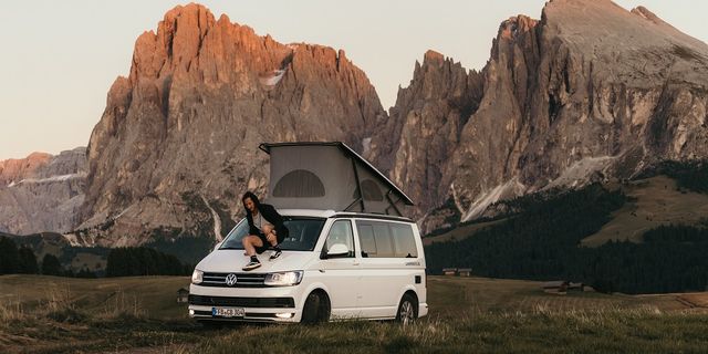 A VW California van is parked in front of some mountains and a man, who was sitting on top of it, is sliding down back onto the ground.