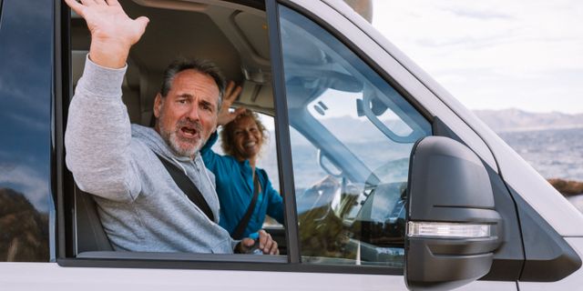Ein Mann winkt aus einem VW Grand California auf dem Beifahrersitz, daneben sitzt eine Frau