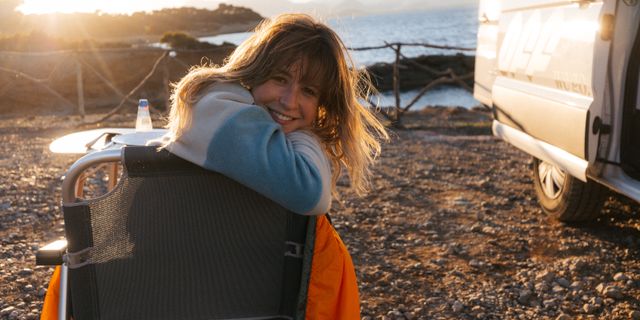 Warme Reiseziele April: Junge Frau sitzt lachend neben einem Camper am Meer in der Sonne