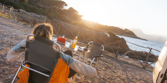 Mit Camping Tipps können Camper-Neulinge entspannt verreisen: Wie die junge Frau, die im Campingstuhl vor einem Tisch in der Sonne aufs Meer schaut und die Arme zur Seite streckt.