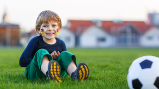 Ein kleiner Junge mit Deutschland Flaggen auf den Wangen sitzt im Gras neben seinem Fußball und lacht in die Kamera