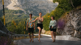 Drei junge Menschen der LGBTQ-Community laufen springend einen Berg auf einer Straße nach oben, im Hintergrund sieht man eine Berglandschaft.
