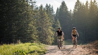Ein Mann und eine Frau mit Fahrradhelm fahren auf Mountainbikes durch ein sonniges Waldstück und lachen dabei
