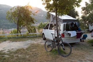 Camping Urlaub mit dem Fahrrad: ein Camper steht auf einem Campingplatz mit toller Aussicht von oben auf eine Stadt, während Wäsche auf dem fahrradträger trocknet und ein Fahrrad an den Baum angelehnt ist