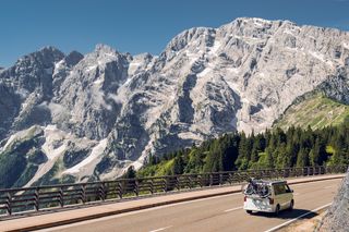 Ein weißer Campervan mit Fahrrädern fährt eine Straße entlang, im Hintergrund sieht man eine Bergkette, Wälder und blauen Himmel