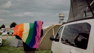 Eine Person mit Regenbogenflagge in der Hand steht neben einem Camper - der Gay Travel Index bewertet, wie LGBTQ-freundlich Länder weltweit sind.