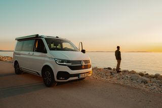 So schön kann Camping in der Nebensaison sein: ein Mann steht vor seinem Wohnmobil am Meer und schaut auf den Horizont, an dem die Sonne untergeht