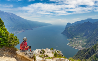 Person in Mountainbikingausrüstung sitzt auf Fels und überblickt den Gardasee von oben