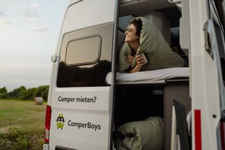ACSI Camping Nebensaison: Frau mit Sonnenbrille liegt im Camper und schaut aus der hinteren Tür in den Himmel