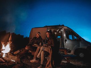 Eine Frau und ein Mann sitzen abends an einem Lagerfeuer vor einem Camper.