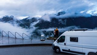 Ein Camper steht auf einer Straße. Zwei Menschen betrachten hinter ihm Berge, die von Nebelschwaden umhüllt sind.