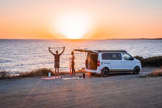 Ein Camper steht an der Küste, im Hintergrund geht die Sonne unter. Zwei Menschen daneben dehnen sich bei ihren Sportmatten.
