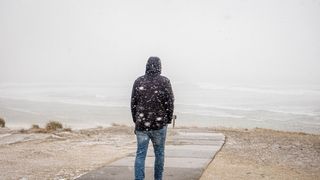 A man is walking on the beach in a dark jacket. There is snow falling around him.