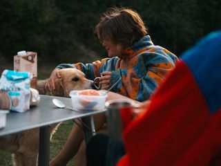 Freunde sitzen beim Frühstück an einem Campingtisch draußen. Eine Frau streichelt einen Hund und hält in der anderen Hand eine Campingtasse.