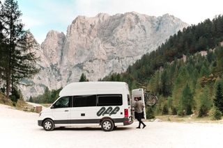 Ein VW Grand California Van steht vor einer Bergkulisse beim Camping in Südtirol. Ein Mann öffnet die Heckklappe.