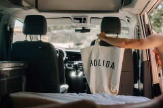 Im Innern eines VW California Ocean Vans hängt ein Mädchen einen Leinenbeutel mit der Aufschrift 'Holiday' über die Kopfstütze des Beifahrersitzes.