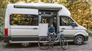 Mann sitzt auf der Treppe eines weißen CamperBoys Camper, der auf einem Campingplatz in Holland steht und lächelt in die Kamera, vor ihm steht sein Fahrrad