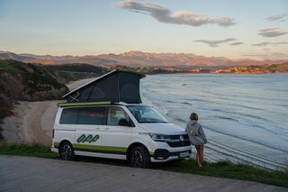 Ein Camper mit Aufstelldach am Straßenrand an der Küste in Portugal. Daneben steht eine junge Frau und schaut aufs Meer, während die Sonne untergeht.