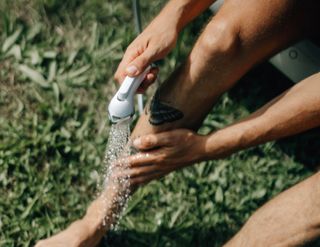 Eine Person wäscht sich die Beine in der Wiese mit dem Duschkopf des Wohnmobils ab: Auch dafür muss man das Trinkwasser im Wohnmobil keimfrei halten und vom Biofilm befreien.