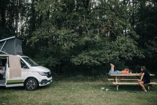 Beim Camping in Estland liegt ein Mädchen auf einem Picknicktisch, ihr Vater sitzt auf der Bank. Daneben steht geparkt ein VW California Ocean Van.