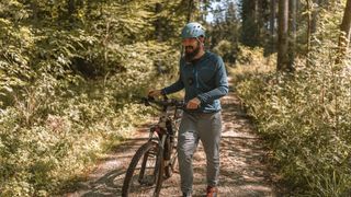 Ein junger Mann mit Helm und Sportjacke bei seinem Urlaub mit dem Fahrrad in Deutschland schiebt sein Mountainbike auf einem Waldweg