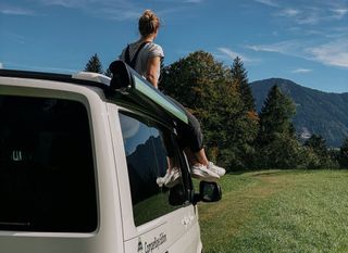 Mädchen sitzt auf dem Dach eines VW California Ocean und blickt in bewaldete Berglandschaft
