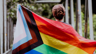 Eine Person, fotografiert von hinten, hält die bunte Regenbogen Flagge der LGBTQIA+-Community in den Händen.