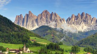 Die Dolomiten haben viele schönste Orte zu bieten: ein Bergmassiv vor blauem Himmel, im Tal sieht man ein idyllisches Dort mit einer Kirche