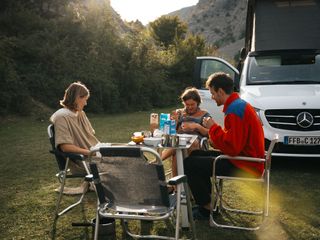 Das Trinkwasser frisch und sauber halten ist auch fürs Kochen wichtig: Eine Freundesgruppe sitzt um ihren Campingtisch herum auf einer sonnigen Wiese neben ihrem Camper. und frühstückt