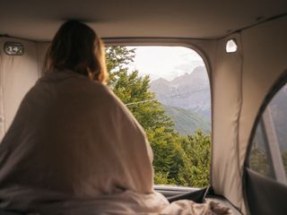 Vorteile Aufstelldach: Eine junge Frau sitzt aufrecht im Aufstelldach ihres Campers und schaut durch das Fenster nach draußen auf die Berglandschaft.