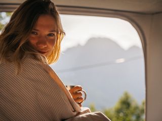 Aufstelldach nachrüsten: Das kann sich lohnen, um so schöne Ausblicke wie diesen zu bestaunen. Hier sitzt eine junge Frau mit Kaffeetasse im Aufstelldach und lächelt in die Kamera.