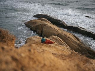 Eine Person sitzt mit einer Decke auf einem Felsen an der Küste und blickt aufs Meer.