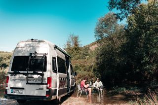 Ein VW Grand California Off Camper und daneben ein Paar, das im seichten See auf Campingstühlen sitzt und isst