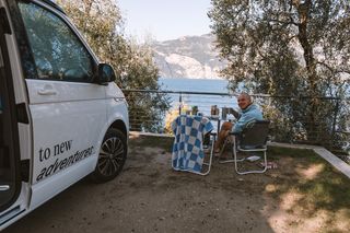 Die Campingplatzwahl ist wichtig beim Camping für Anfänger: Ein junger Mann in blauem Pulli sitzt auf dem Campingplatz neben seinem Camper und frühstückt mit Blick auf den Gardasee.