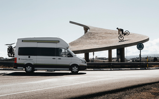 VW Grand California vor einem Bikepark mit einem Mountainbiker.