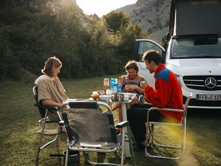 Eine Freundesgruppe sitzt um einen Campingtisch beim Frühstück beim Camping im Münstertal. Im Hintergrund ein Mercedes Marco Polo Camper.