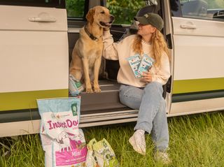 Eine Frau und ein Hund sitzen im Inneren eines Campers an der Schiebetür. Neben dem Camper und in der Hand der Frau ist Hundefutter von Green Petfood zu sehen.