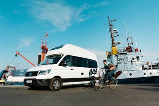 Ein VW Grand California Van steht geparkt an einem Hafen, ein Mann fährt davor Fahrrad.